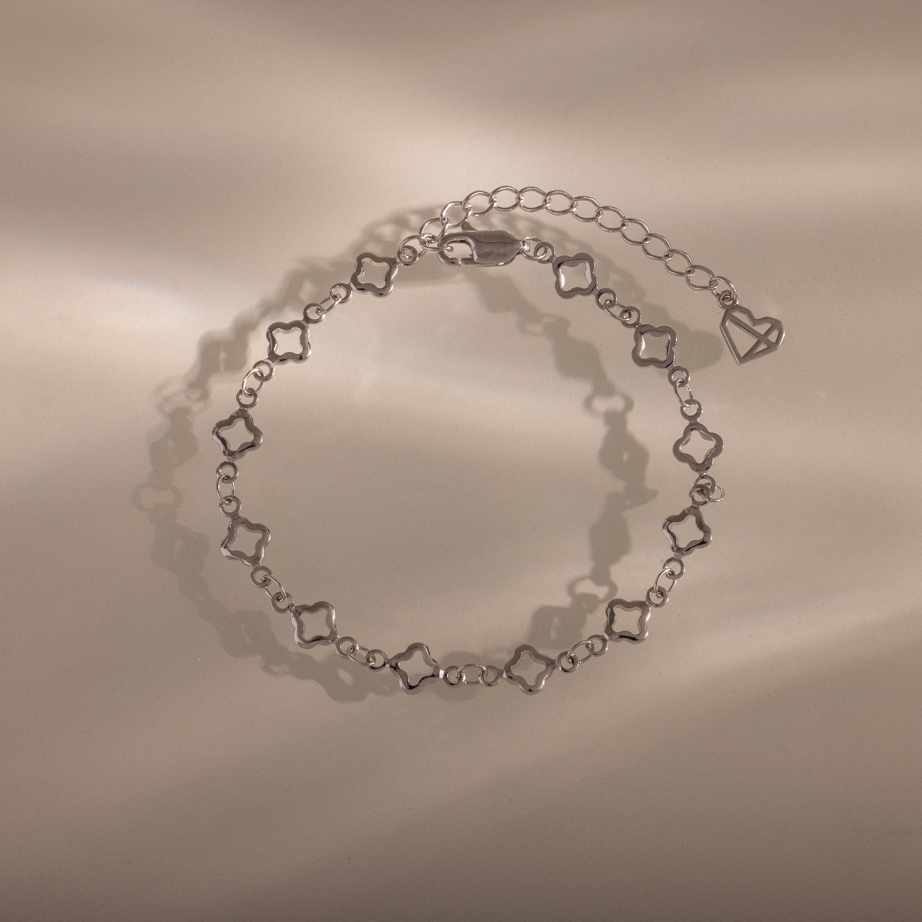 The Clover Station Bracelet in Silver features delicate clover-shaped links and a heart charm, elegantly displayed on a beige surface.