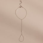 Minimalist silver hand chain bracelet with delicate links and a teardrop-shaped loop, laid flat against a beige background.