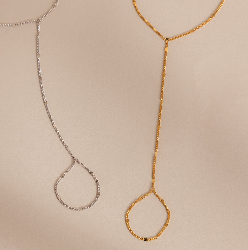 Gold and silver curb hand chain bracelets with finger loops displayed flat on a neutral beige background, showcasing both color options.