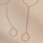 Gold and silver curb hand chain bracelets with finger loops displayed flat on a neutral beige background, showcasing both color options.