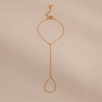 Minimalist gold hand chain bracelet with delicate links and a teardrop-shaped loop, laid flat against a beige background.