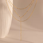 The Sparkling Layered Lariat Necklace in 18K Gold features delicate chains, perfect for effortless layering and summer style, shown against a neutral background with a textured vase.