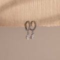 Diamond Drop Huggies in Sterling Silver feature teardrop-shaped crystals hanging from silver hoops, displayed on textured beige paper—an elegant accessory to elevate any semi-formal look against a neutral background.