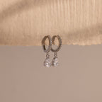 Diamond Drop Huggies in Sterling Silver feature teardrop-shaped crystals hanging from silver hoops, displayed on textured beige paper—an elegant accessory to elevate any semi-formal look against a neutral background.