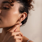 A woman with curly hair pulled back touches her Pave Birthstone Drop Huggies, her face partially visible.