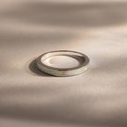 The White Opal Inlay Band in Sterling Silver showcases a shimmering opal inlay set on a sleek silver band, displayed on a beige surface with soft shadows for elegant gemstone appeal.