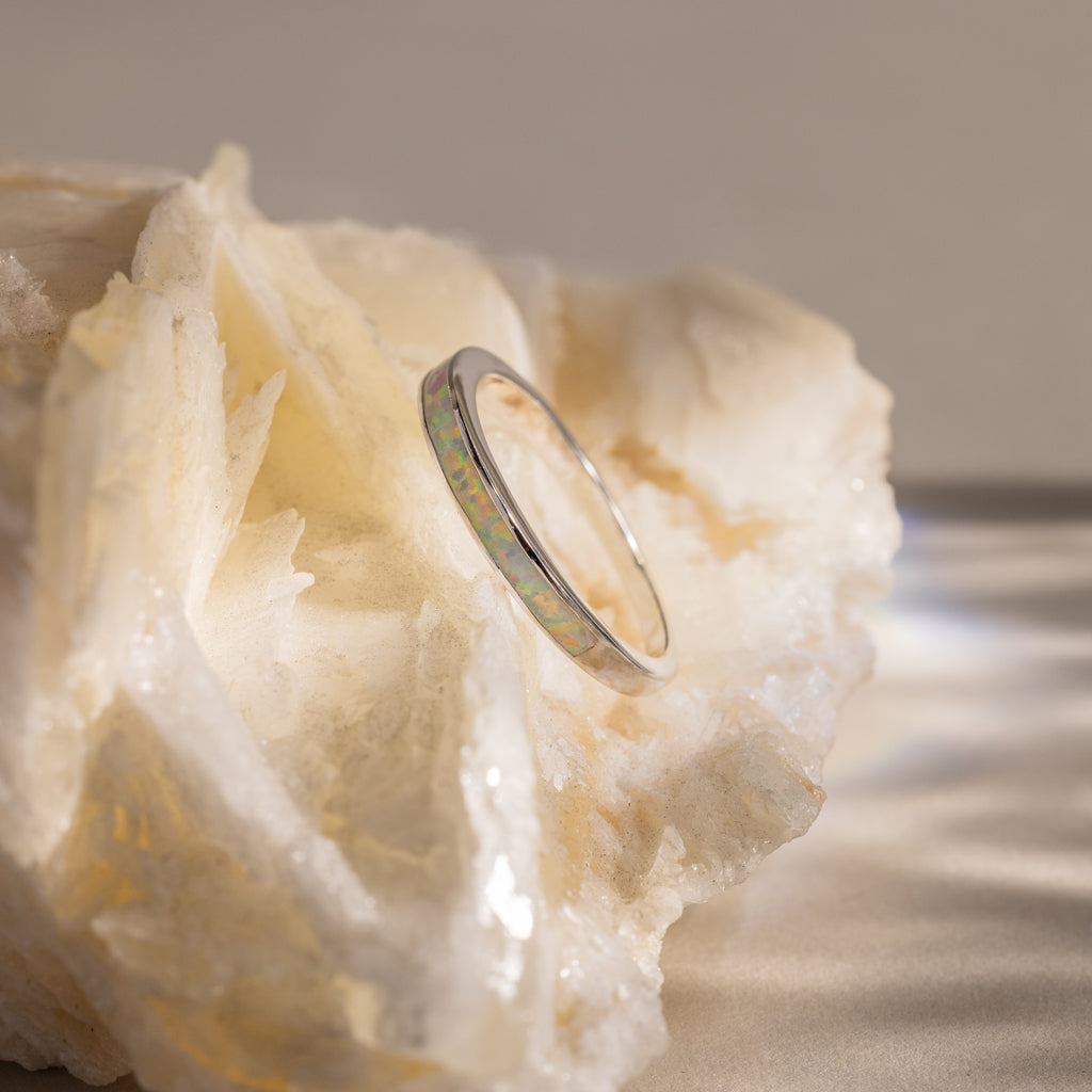 The White Opal Inlay Band in Sterling Silver rests on a rough, pale crystal surface, its iridescent sheen capturing the ethereal beauty of gemstone jewelry.