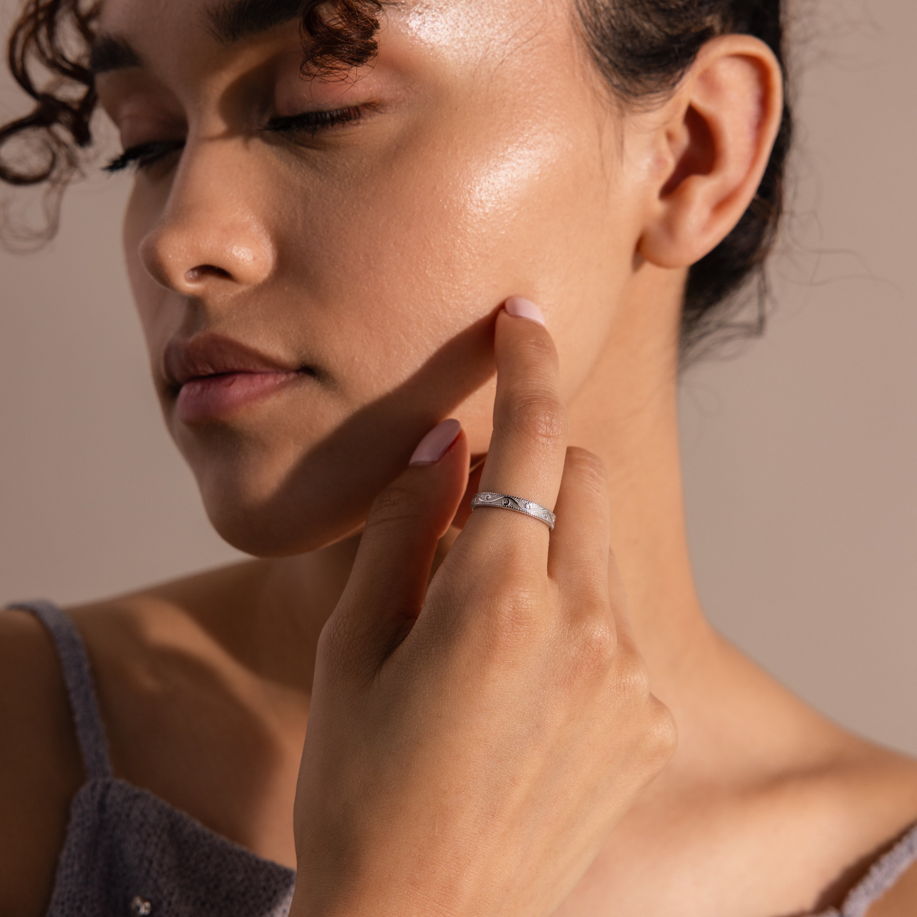 Woman with curly hair touching her cheek, eyes closed, in soft lighting, wearing the Filigree Ring in Sterling Silver with an engraved band.