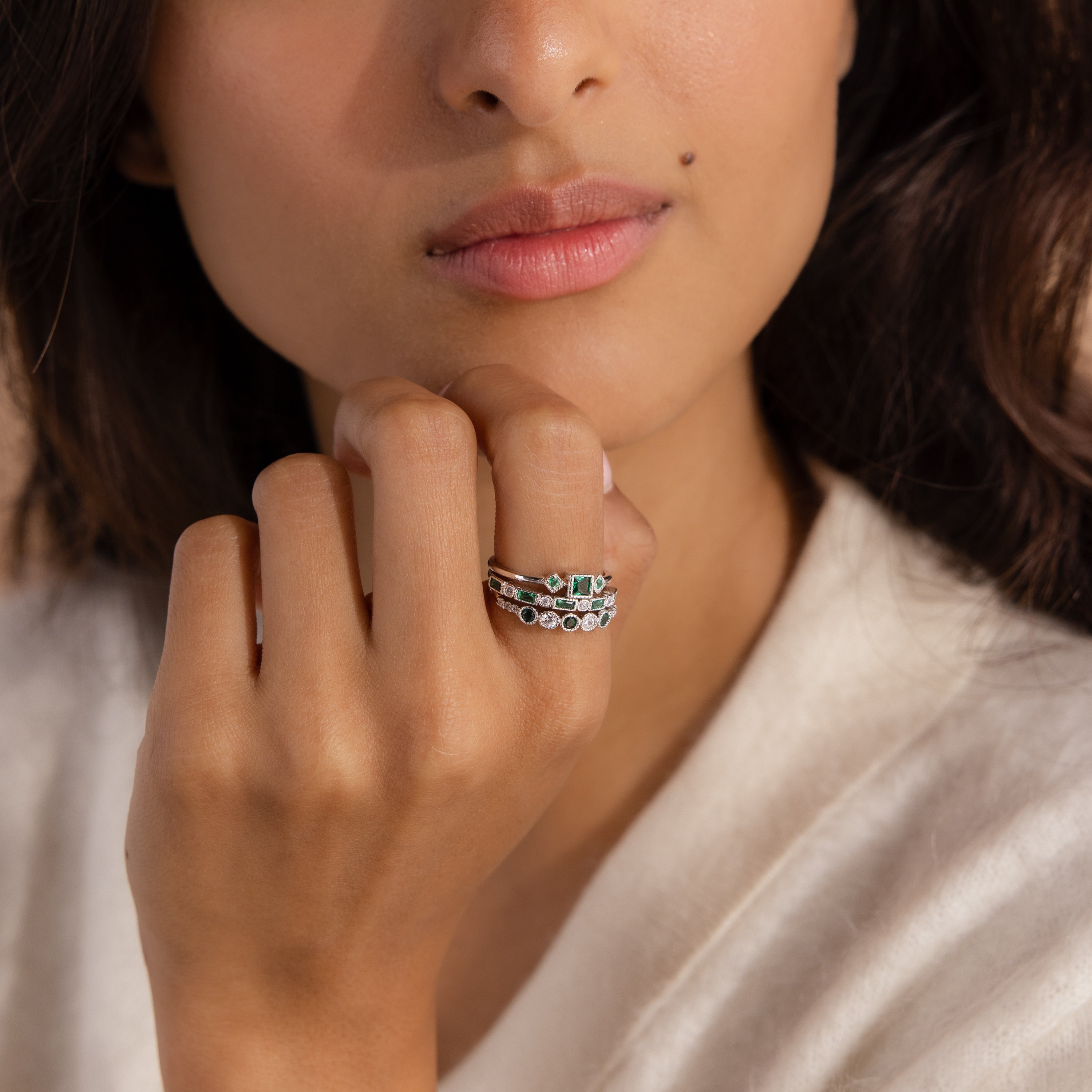 A woman with medium skin tone wears the Emerald Stacking Ring Set in Sterling Silver, along with other silver rings, gently touching her chin in soft lighting.