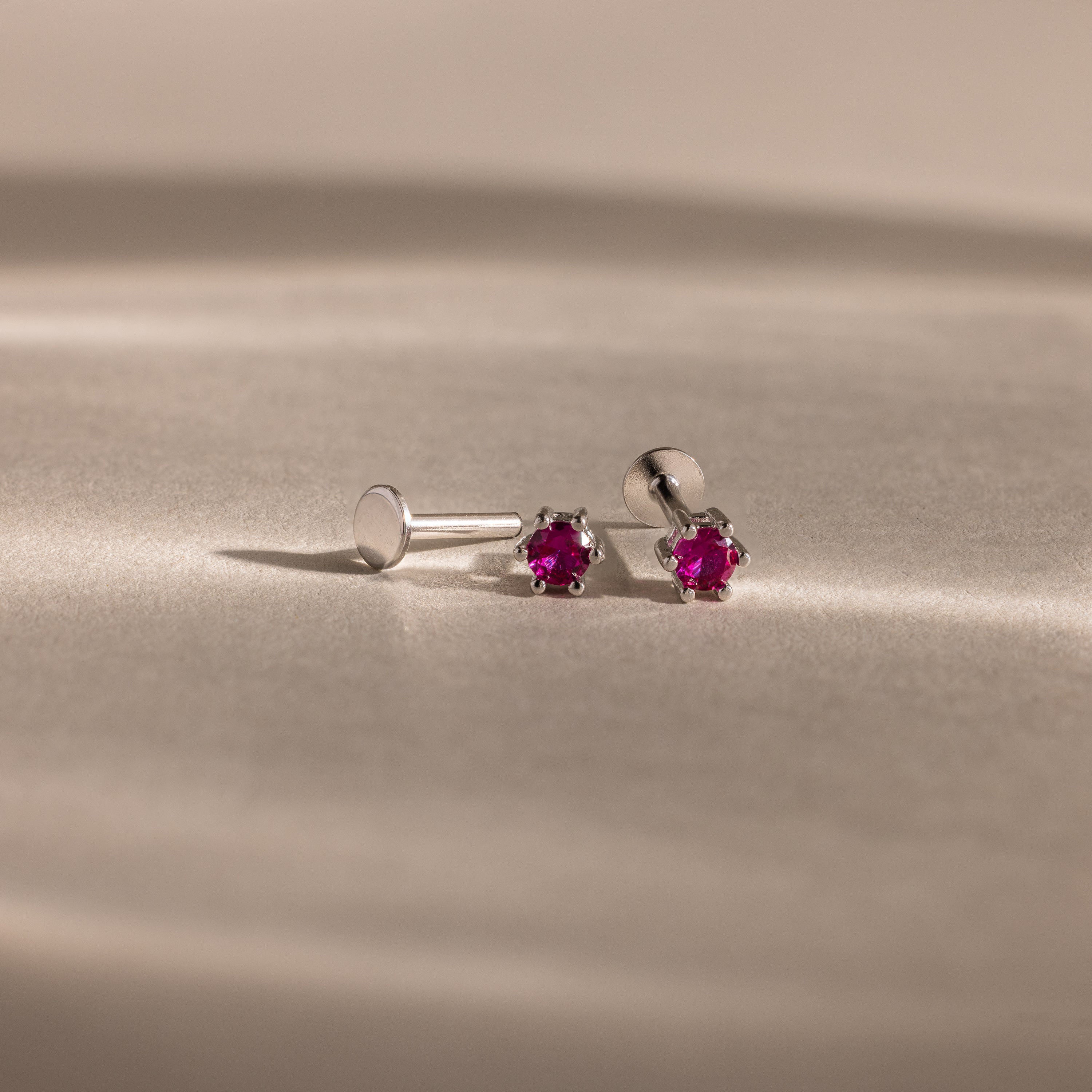 Ruby Flatback Studs—silver flatback earrings with small round red gemstones—make elegant birthstone jewelry, beautifully displayed on a soft beige surface.