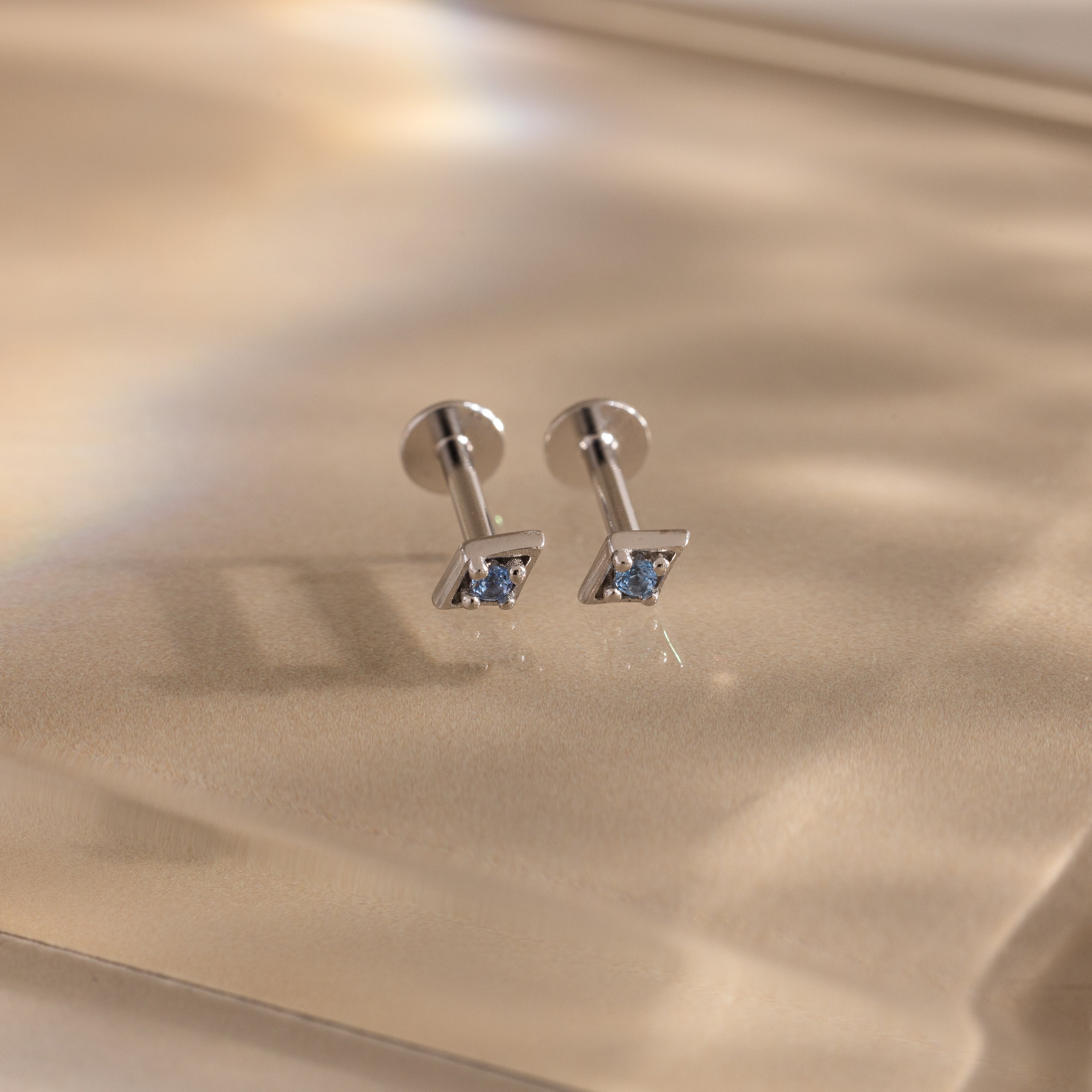 Pair of silver geometric flat back aquamarine stud earrings on a reflective gold surface