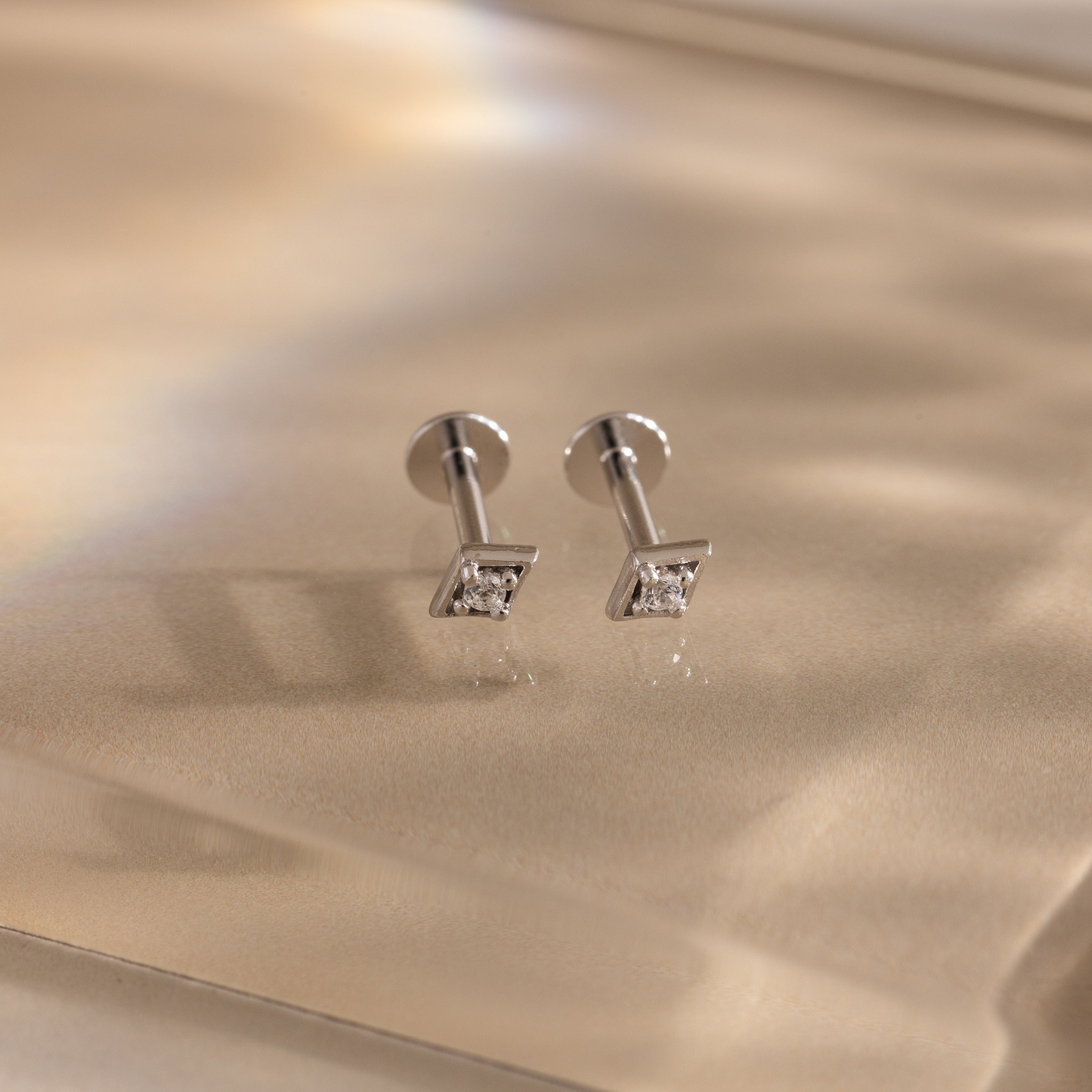 Pair of silver geometric flat back diamond stud earrings on a reflective gold surface