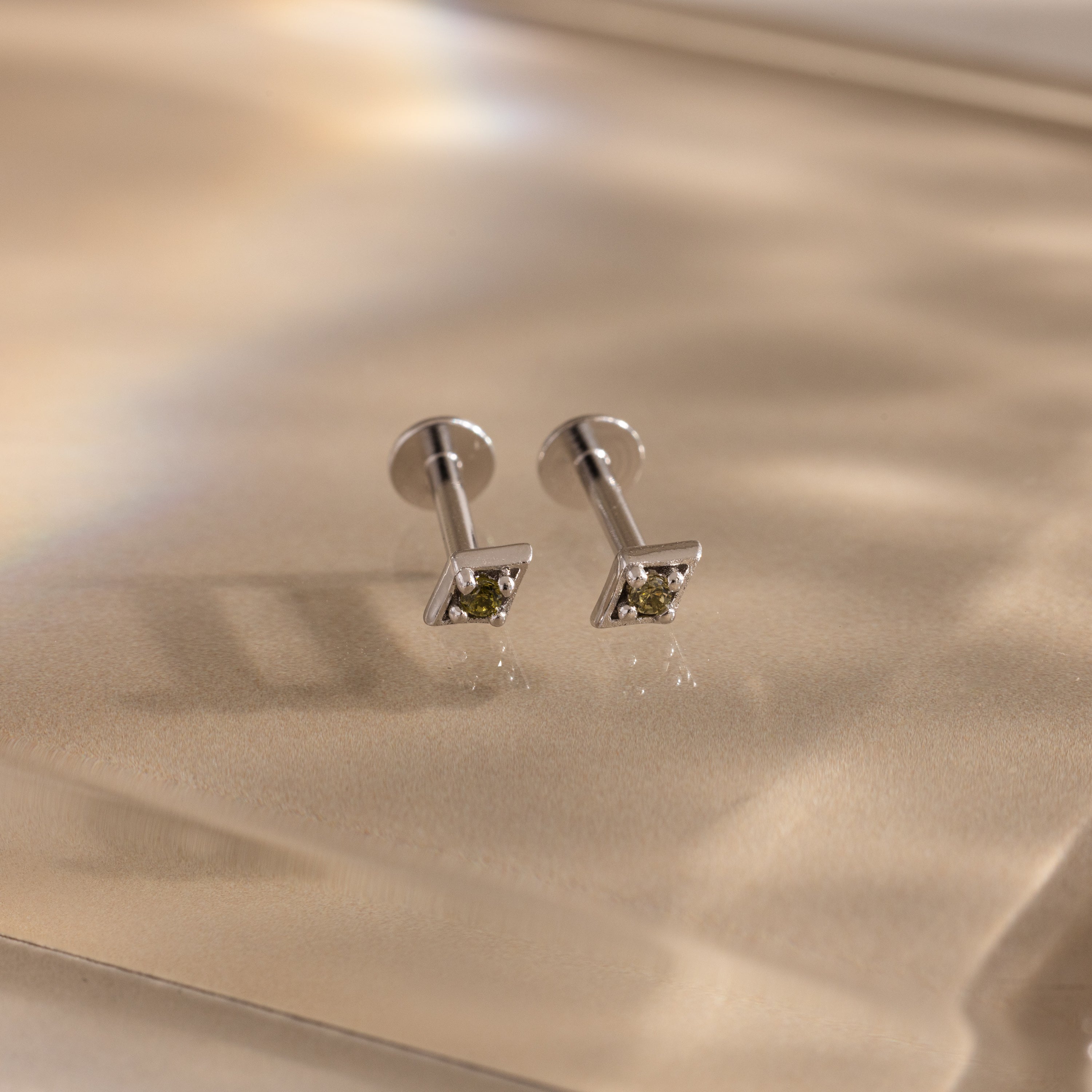 Pair of silver geometric flat back peridot stud earrings on a beige fabric background