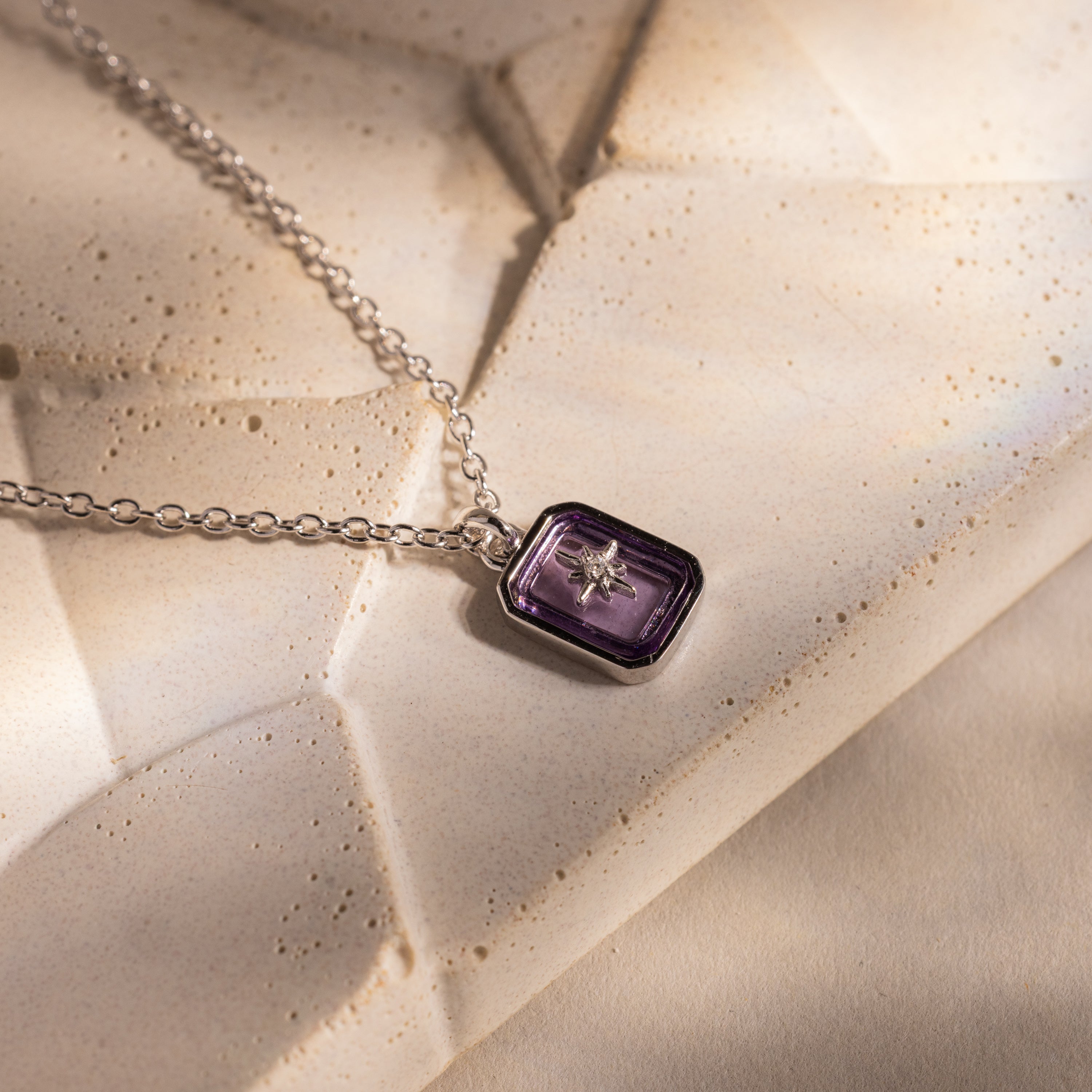The North Star Amethyst Necklace, featuring a rectangular purple crystal pendant on a silver chain, rests on a textured beige surface.
