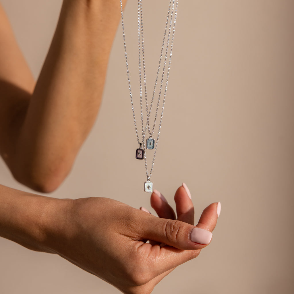 A hand displays three silver necklaces with rectangular gemstone pendants, featuring the sparkling North Star Aquamarine Necklace, against a neutral background.