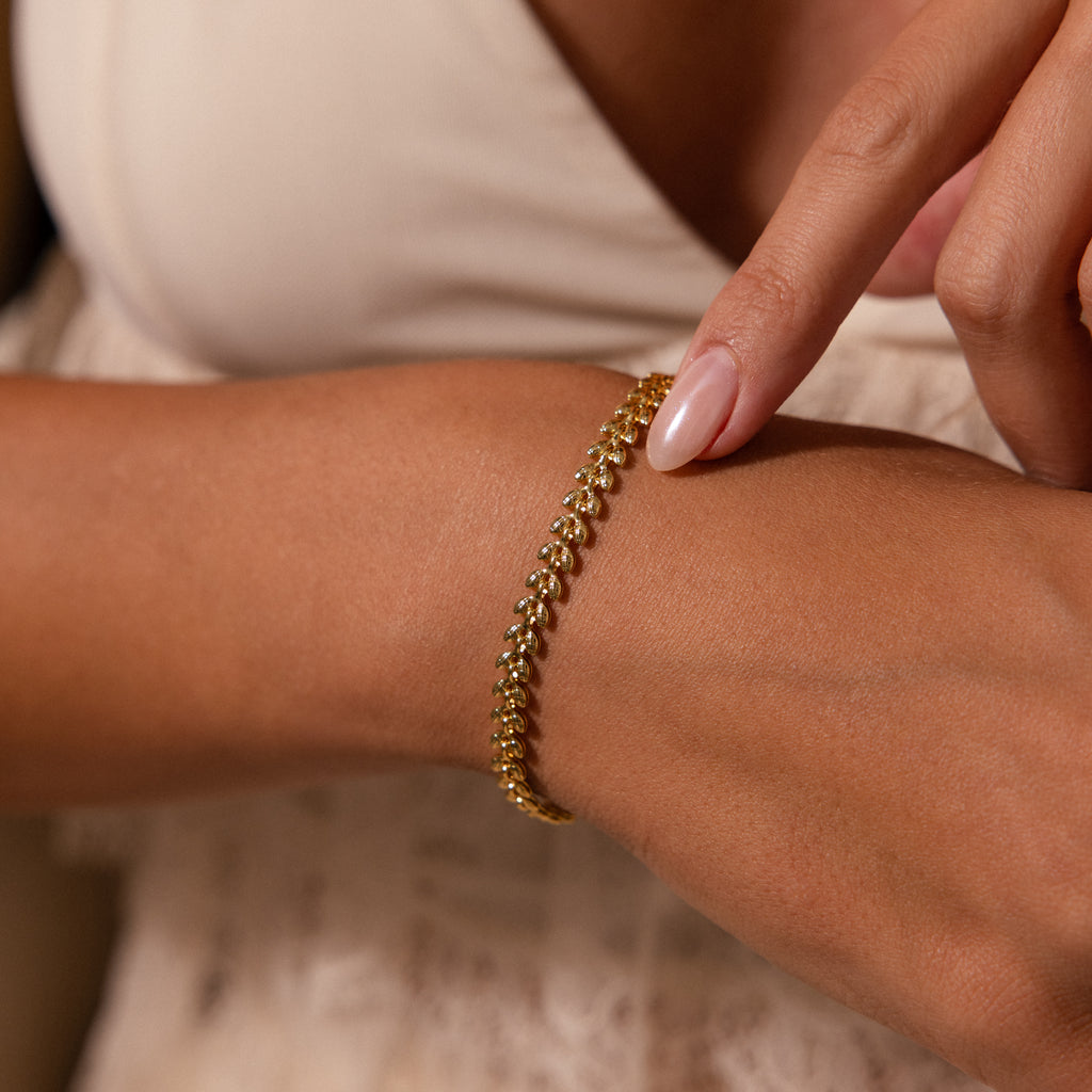 A person points to a Fishtail Chain Bracelet in 18K Gold on their wrist, displaying its detailed interwoven links while wearing a cream-colored top.