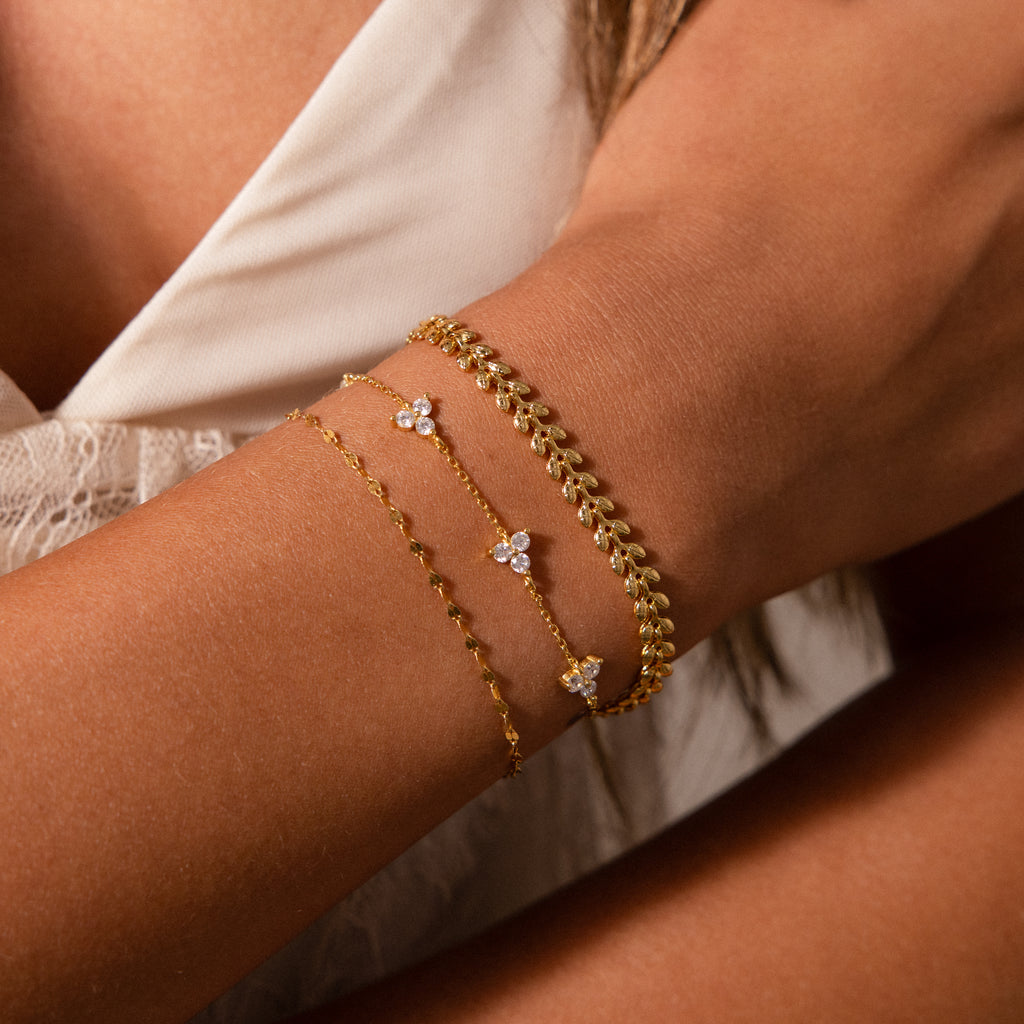 A woman’s arm adorned with three delicate gold bracelets, including the Fishtail Chain Bracelet in 18K Gold, each featuring small gemstones, against a white lace top.