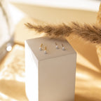 Four Dainty Pearl Studs in Sterling Silver are displayed on a white rectangular block with dried grass in the background, accentuating their delicate elegance.