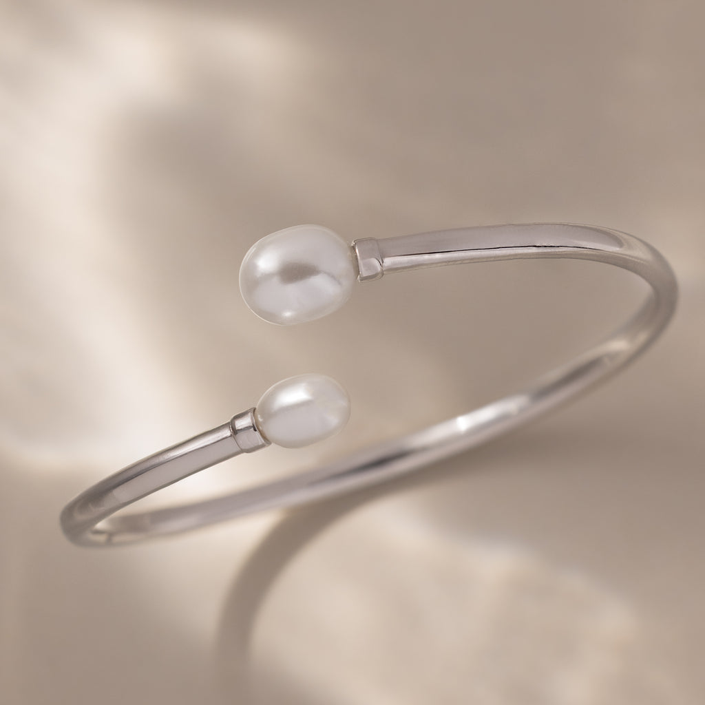 The Serenity Pearl Cuff Bracelet in Silver features radiant pearls on each open end, elegantly displayed against a soft beige background.