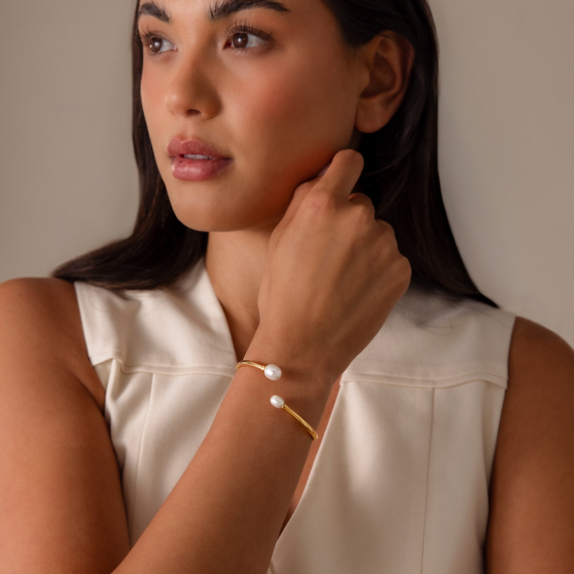 Woman in a sleeveless cream top wears the Serenity Pearl Cuff Bracelet in 18K Gold, featuring a modern open band with two pearls, while looking to the side.