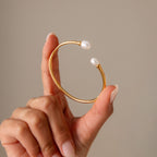 A hand displays the Serenity Pearl Cuff Bracelet in 18K Gold—an open band with pearls on each end—against a neutral background.