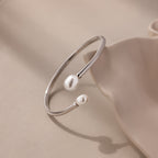 The Serenity Pearl Cuff Bracelet in Silver features an open band with two luminous pearls, elegantly displayed on a soft beige surface.