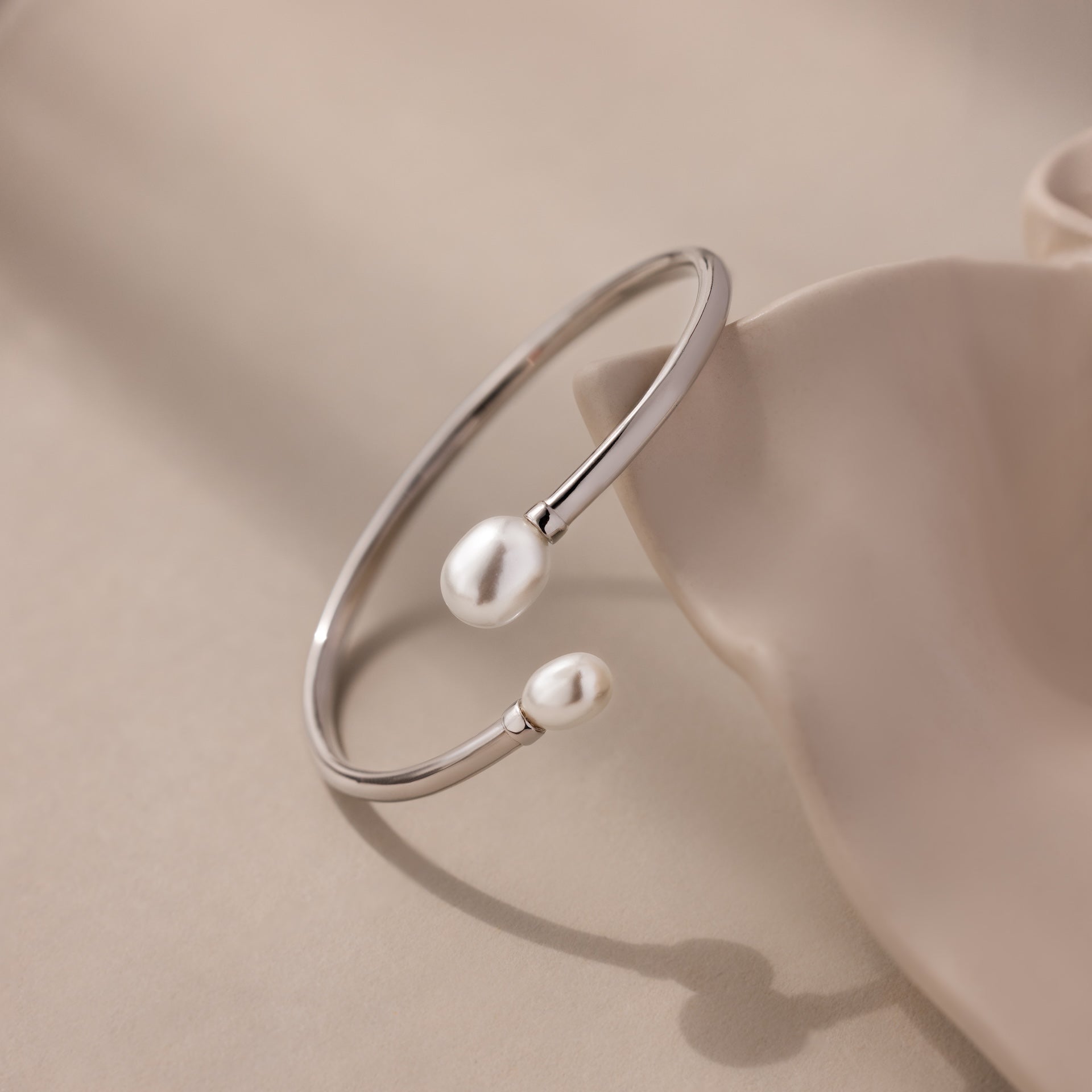 The Serenity Pearl Cuff Bracelet in Silver features an open band with two luminous pearls, elegantly displayed on a soft beige surface.