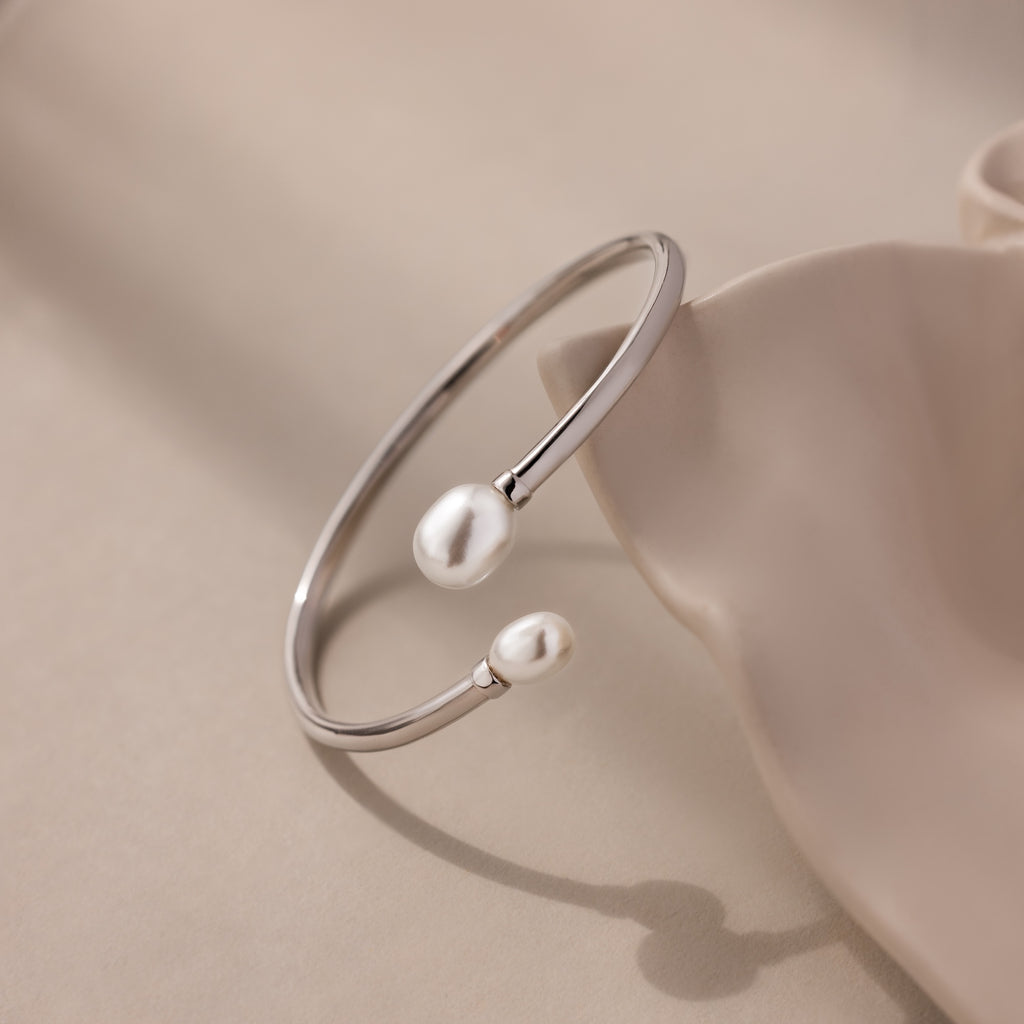 Serenity Pearl Cuff Bracelet: A silver open band bracelet featuring two white pearls, showcased on a soft beige surface.