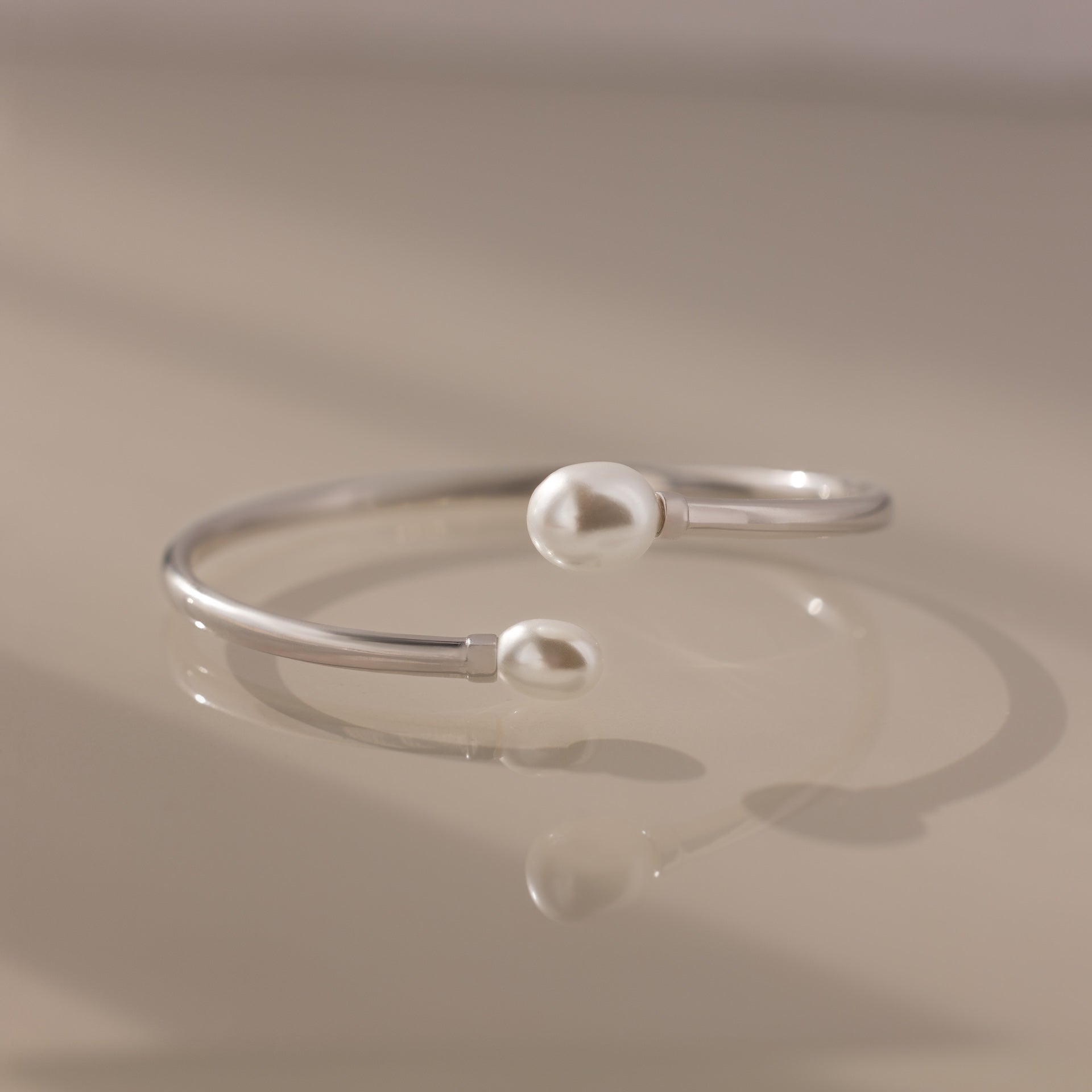 The Serenity Pearl Cuff Bracelet in Silver, featuring luminous pearls at each end, rests gracefully on a beige surface.
