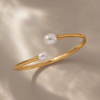 The Serenity Pearl Cuff Bracelet in 18K Gold features an open band with two white pearls on each end, displayed against a soft beige background—a stunning modern pearl accessory.