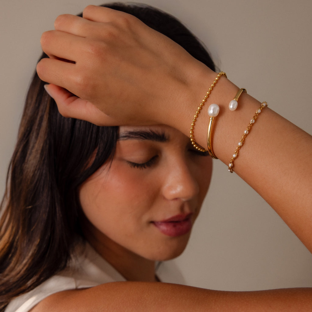 Woman with closed eyes showcases a modern look, wearing the Serenity Pearl Cuff Bracelet—an elegant gold and pearl open band—on her wrist against a neutral background.