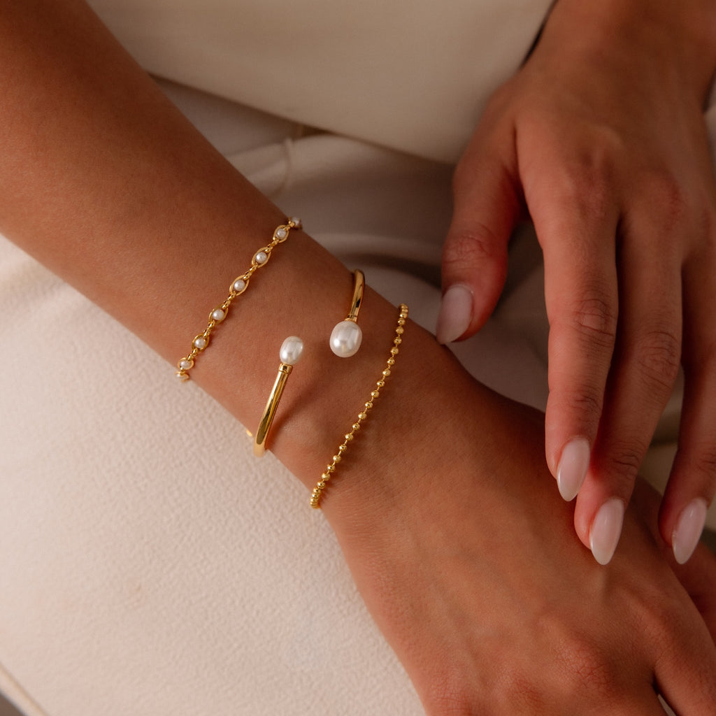 A close-up of a womans wrist adorned with the Dune Beaded Bracelet in 18K Gold, featuring polished beads and delicate pearls among three elegant gold bracelets.