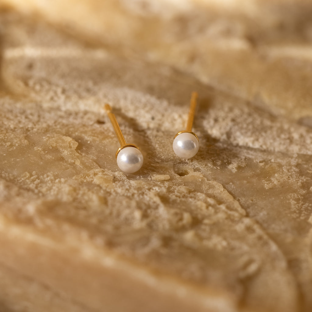 Dainty Pearl Studs featuring round white pearls on a textured beige stone surface—perfect minimalist earrings or for stacking with other styles.