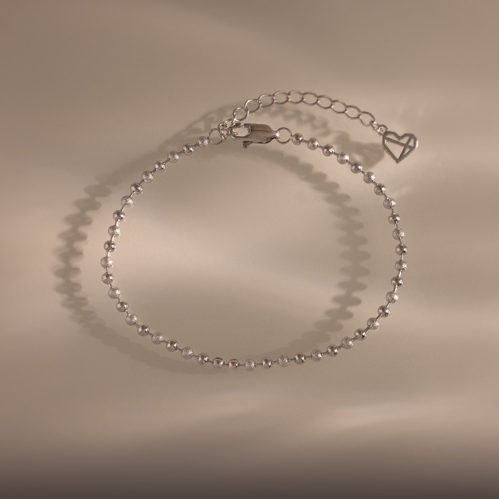 Dune Beaded Bracelet in Silver features polished silver beads, a heart charm, and an adjustable chain, displayed on a neutral background.