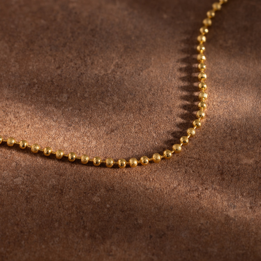 The Dune Beaded Bracelet in 18K Gold, featuring polished beads, is displayed on a brown textured surface.