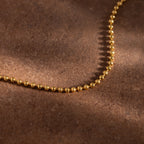 The Dune Beaded Bracelet in 18K Gold, featuring polished beads, is displayed on a brown textured surface.