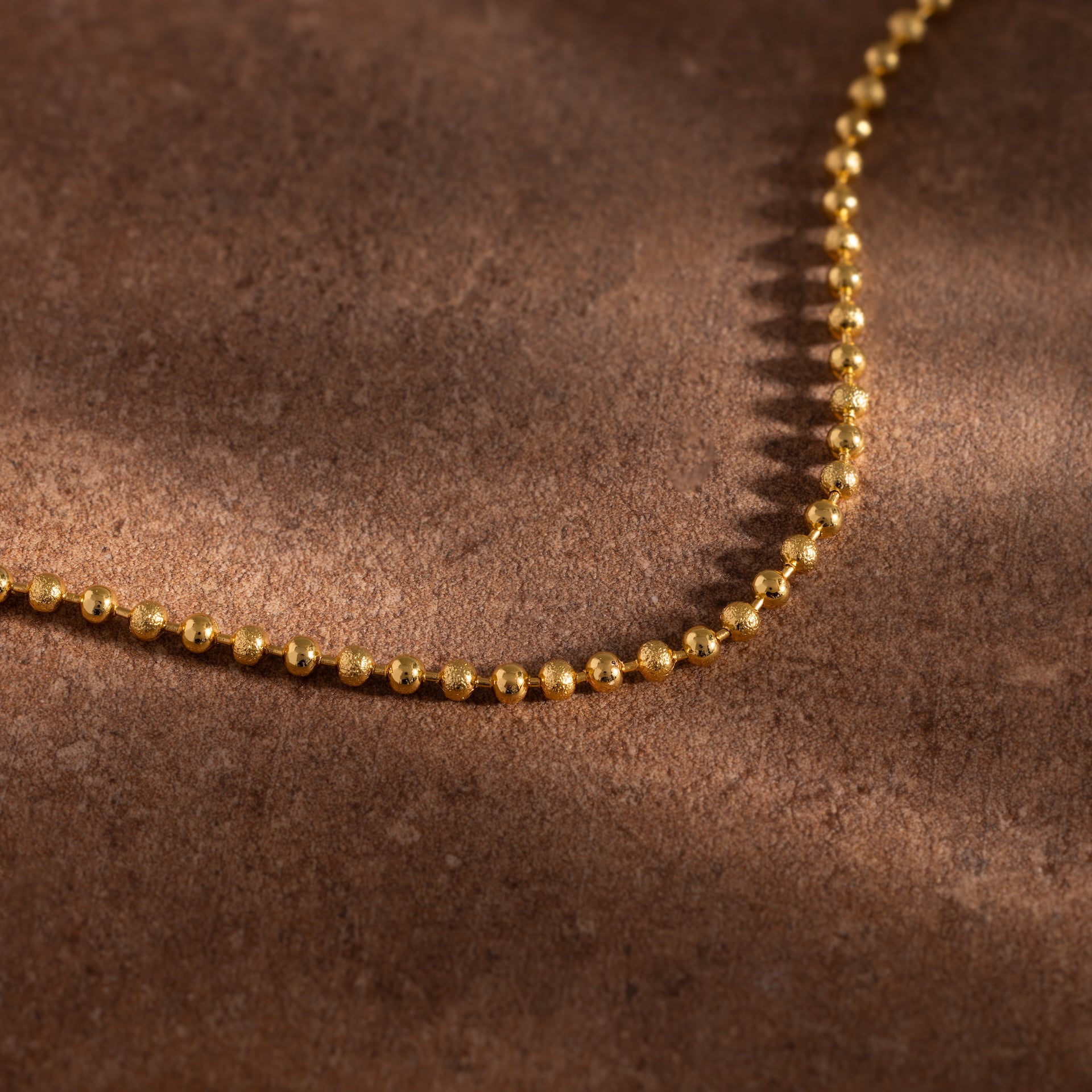 The Dune Beaded Bracelet in 18K Gold, featuring polished beads, is displayed on a brown textured surface.