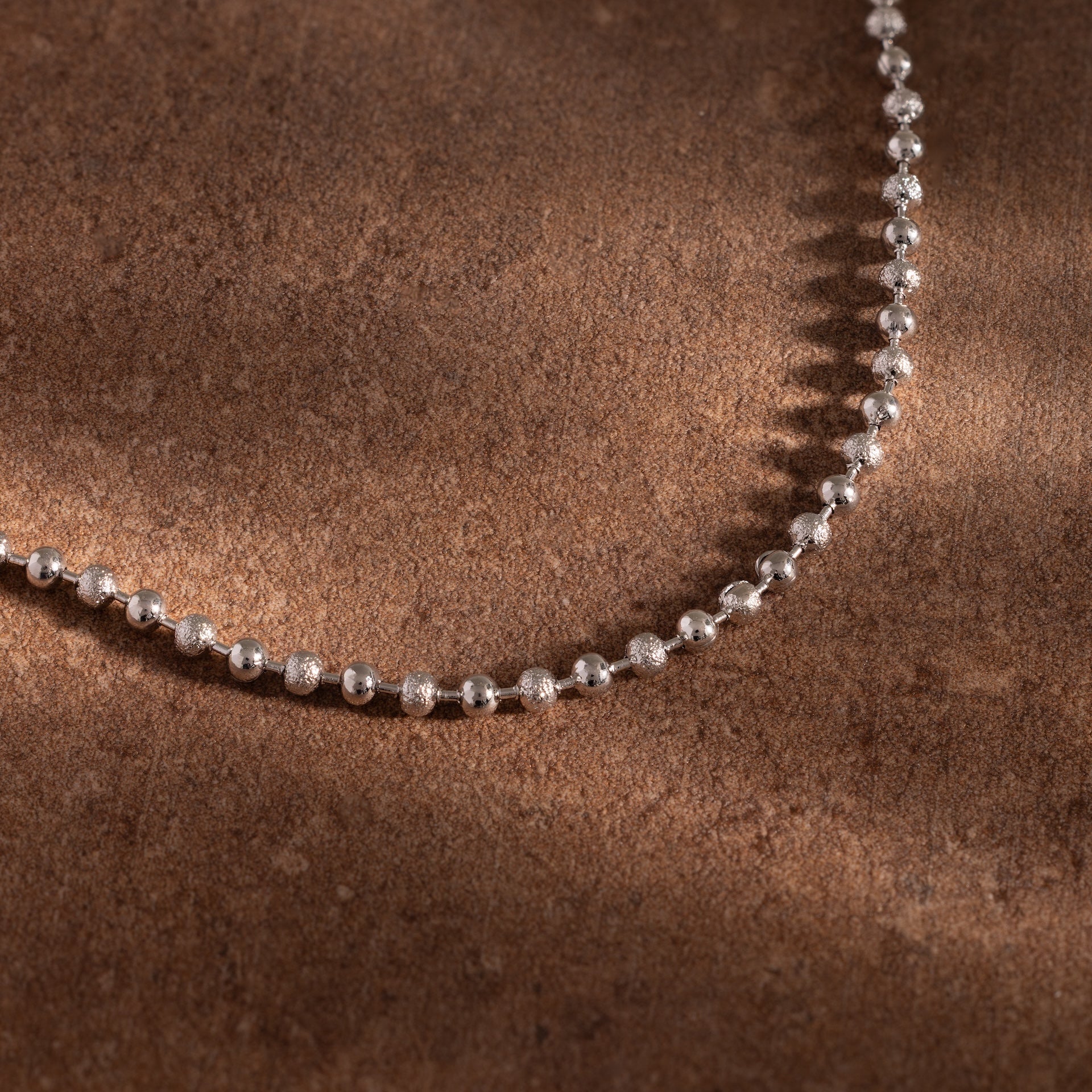 The Dune Beaded Bracelet in Silver rests on a textured brown surface, highlighted by soft shadows and warm lighting.