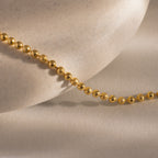 The Dune Beaded Bracelet in 18K Gold features polished beads elegantly draped over a smooth, light stone surface, casting gentle shadows.