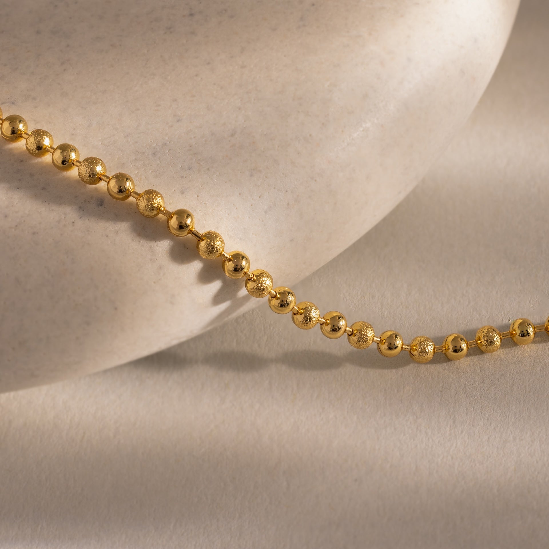 The Dune Beaded Bracelet in 18K Gold features polished beads elegantly draped over a smooth, light stone surface, casting gentle shadows.
