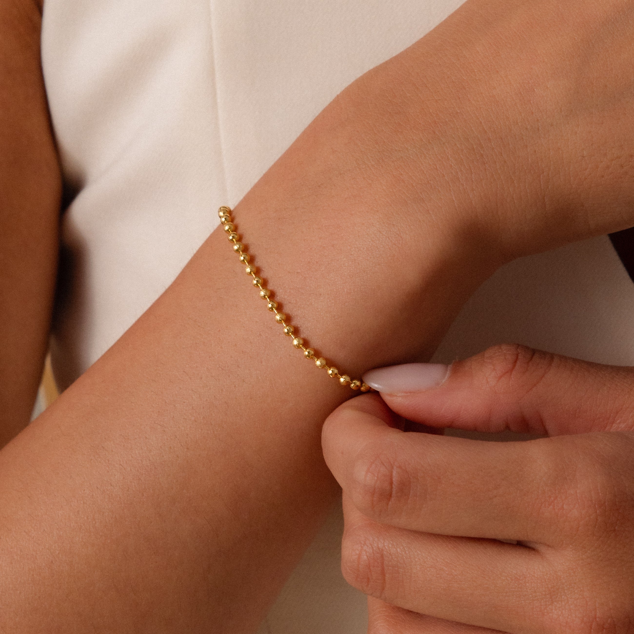 A person clasps the Dune Beaded Bracelet in 18K Gold on their wrist, pairing its polished beads with a light-colored outfit.