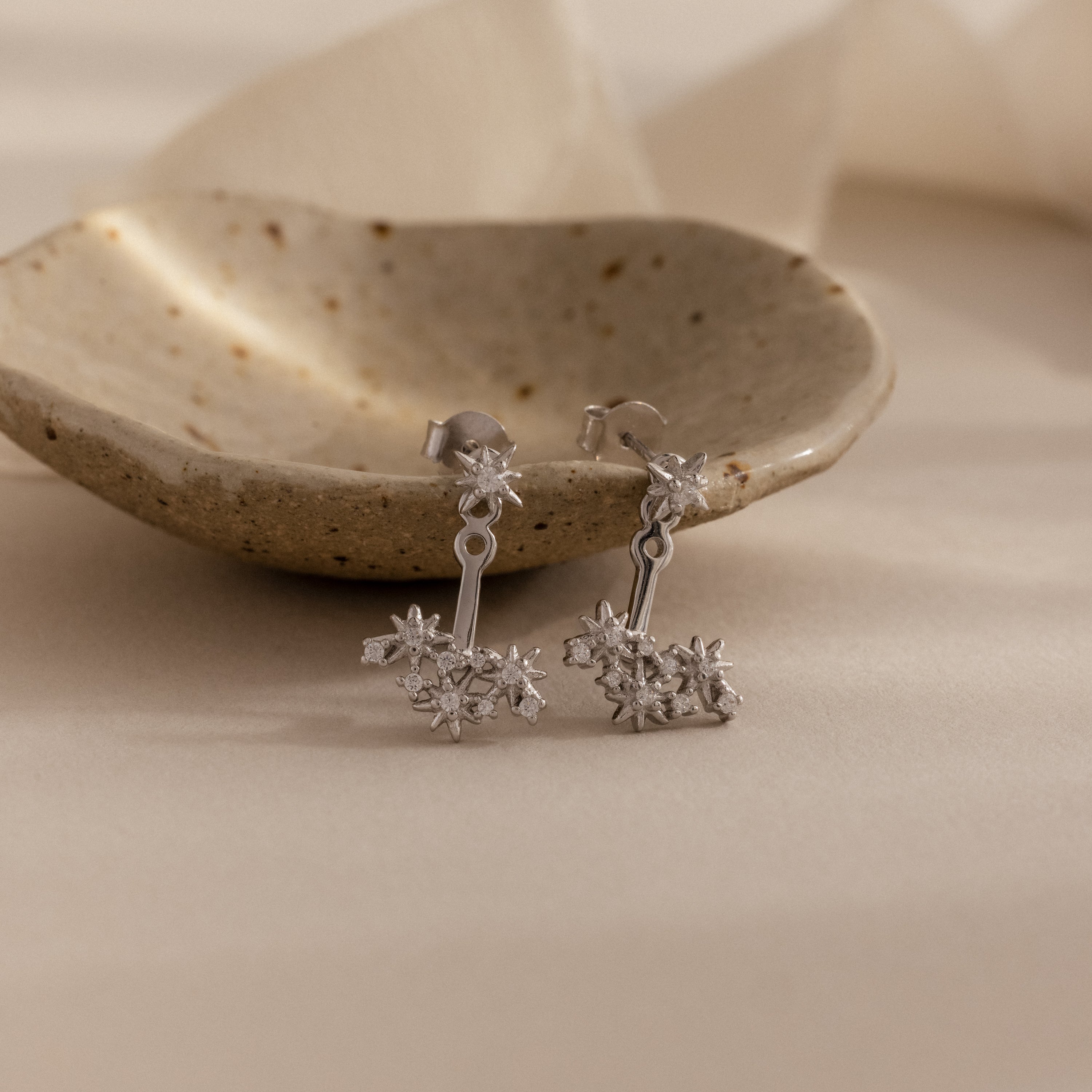 Starburst Cluster Ear Jackets in Sterling Silver, inspired by constellation designs, are displayed on a speckled ceramic dish with a soft beige background and gentle lighting.