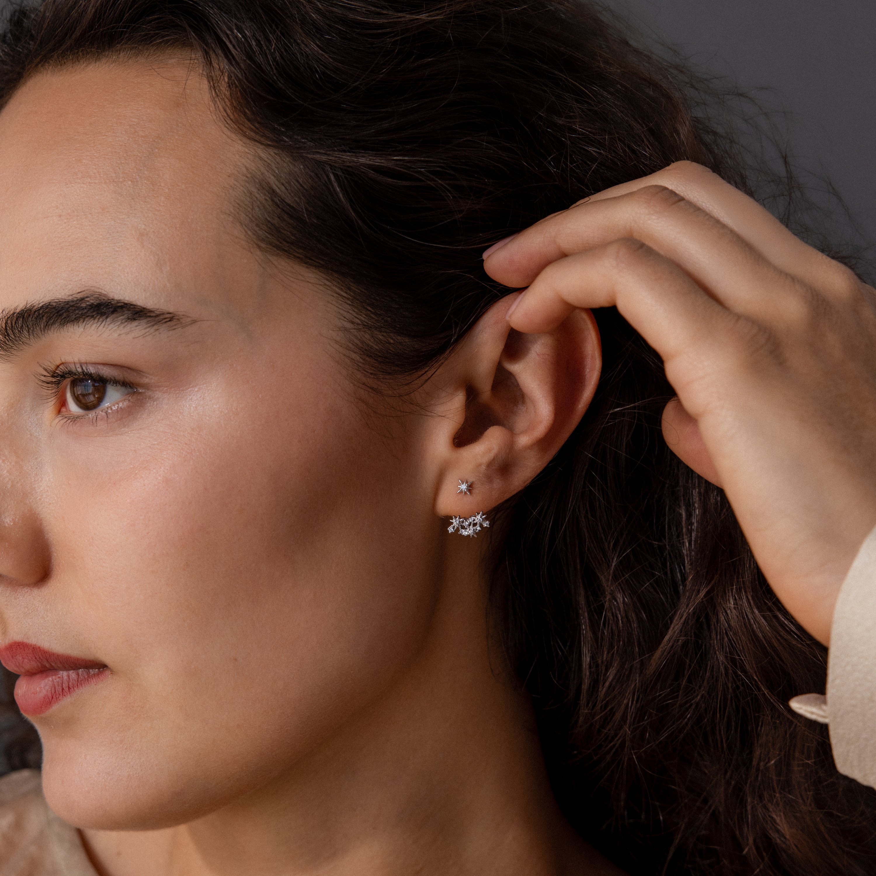 A woman touches her dark hair while wearing two small, delicate Starburst Cluster Ear Jackets in Sterling Silver in her left ear.