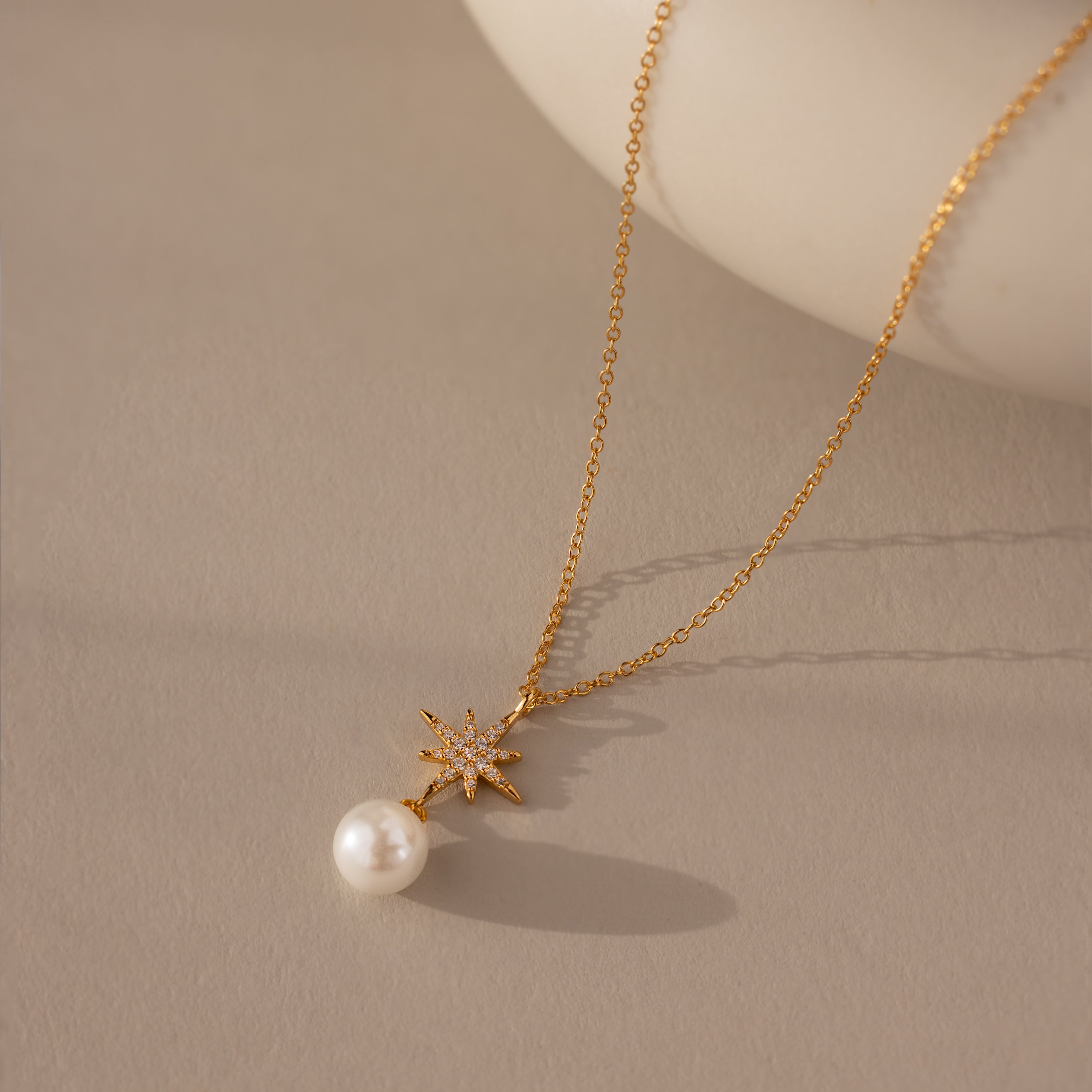 Introducing the North Star Pearl Necklace in 18K Gold, featuring a delicate pearl pendant—ideal for lovers of timeless pearl jewelry. Shown on a neutral beige surface for an elegant touch.