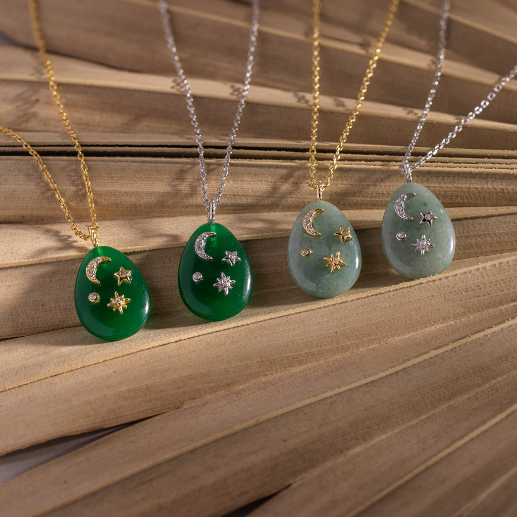 Four Celestial Gemstone Pendant Necklaces with moon and star designs, featuring gold and silver chains and a standout green gemstone piece, are beautifully displayed on a wooden surface.