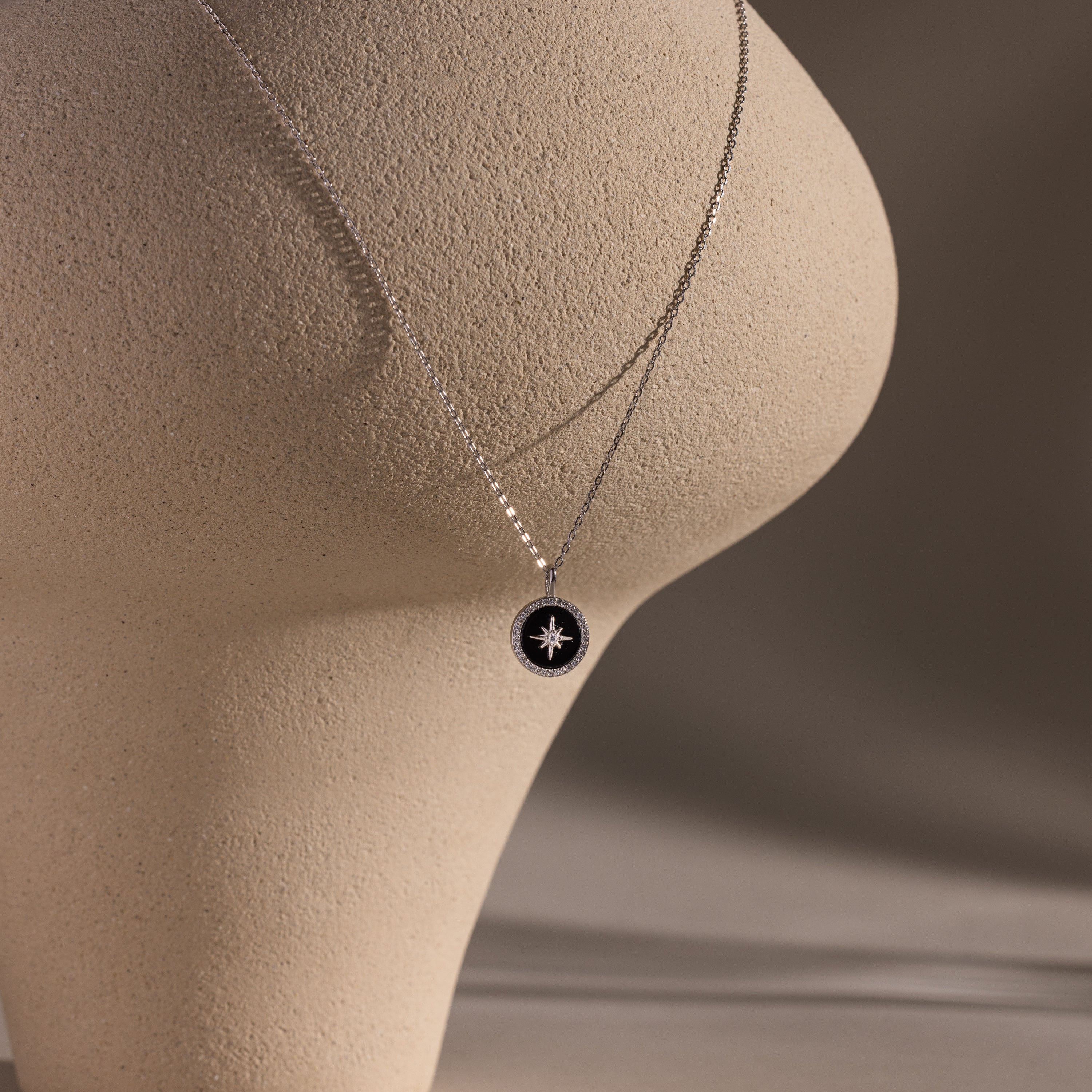 The Onyx Star Pendant Necklace in Sterling Silver is draped over a textured beige abstract sculpture, set against a neutral background.