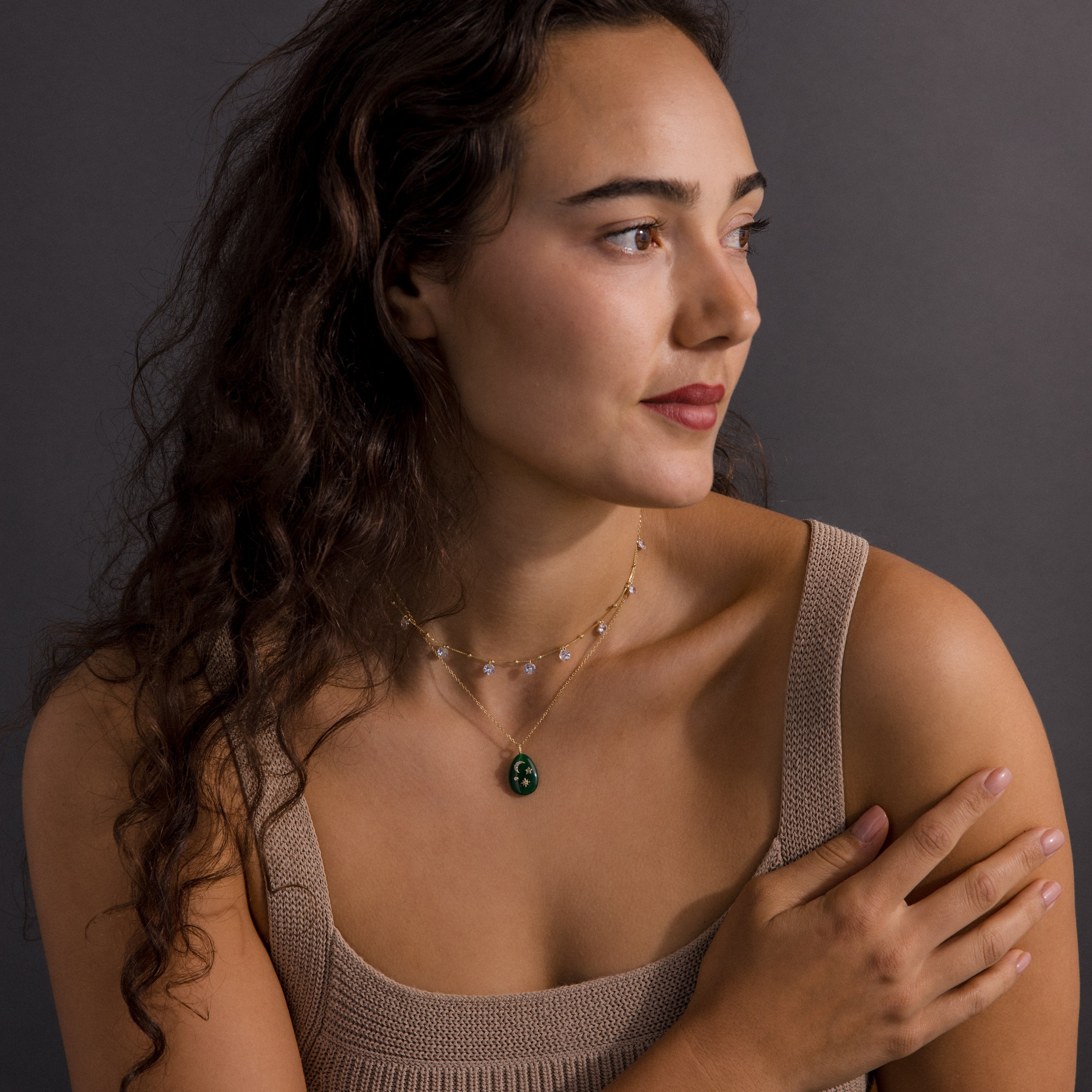Woman with long wavy hair wearing a beige top and layered Celestial Gemstone Pendant Necklaces, gazing to the side.