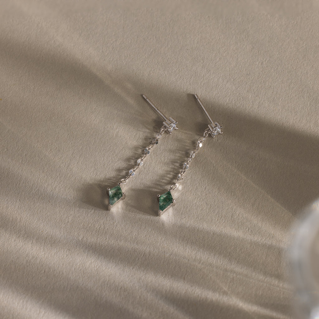 Agate Diamond Dangle Earrings with clear stones and green gemstone ends lie on a textured beige surface—a modern essential jewelry piece.