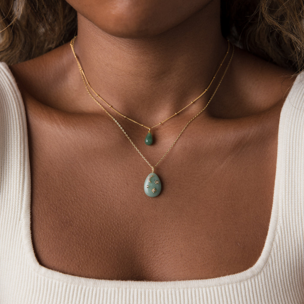 Close-up of a woman wearing two gold necklaces with green stone pendants, including the Celestial Gemstone Pendant Necklace, over a cream ribbed top.