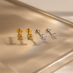 Two pairs of Art Deco flatback stud earrings—one in gold and one in silver—featuring baguette-cut diamonds, displayed on a reflective, warm-toned surface.